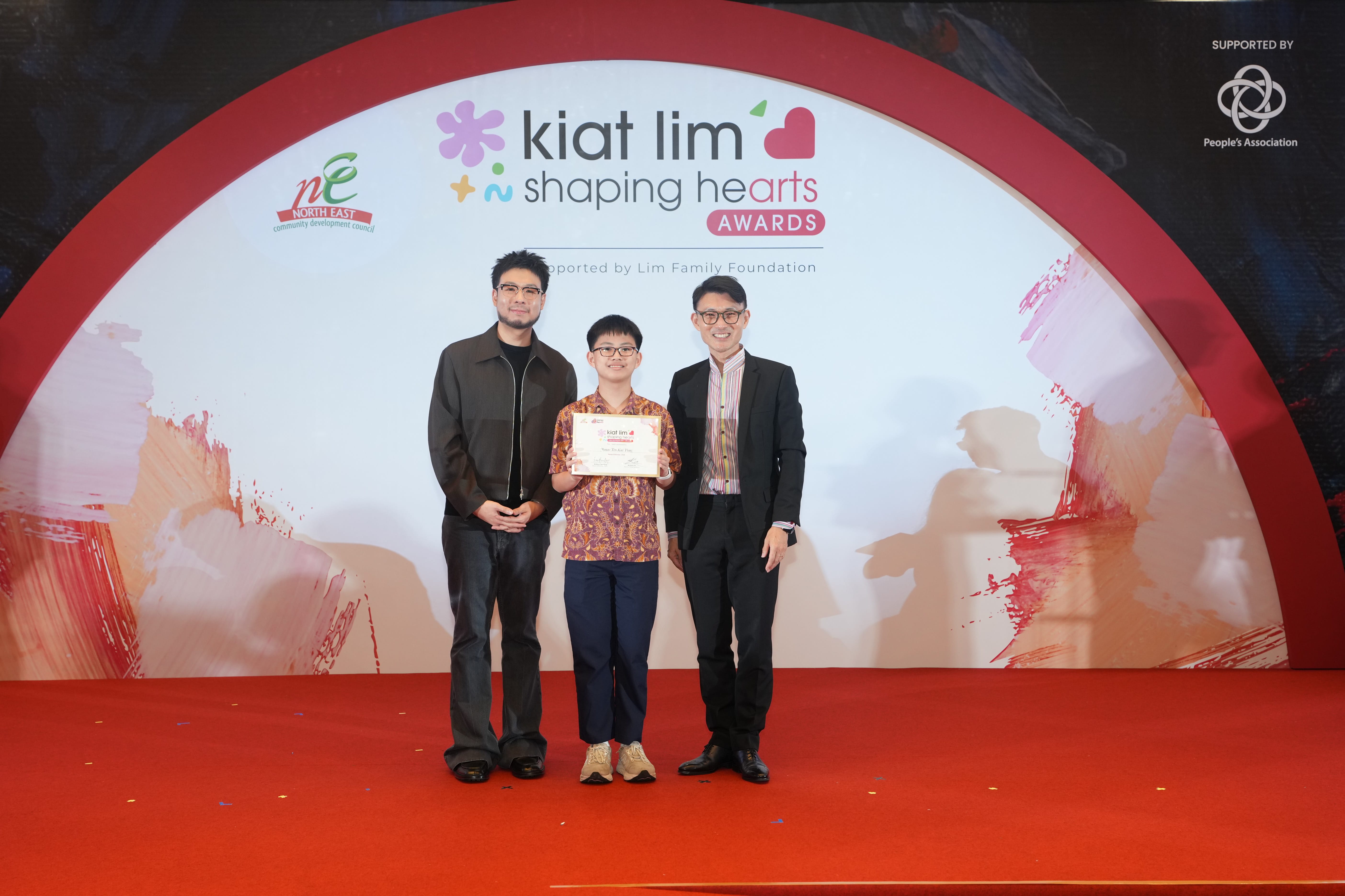 Award recipient standing on stage with the presenter, holding their certificate and posing for a group photo during the award ceremony, with a backdrop reading ‘Kiat Lim-Shaping Hearts Awards'.